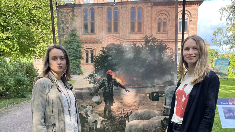 Pinar and Lina standing next to a huge photo of a man saving his cattle from a wild fire. (Photo)
