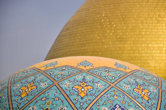 A photo of two domes on top of a shrine and mosque, one in gold and one in blue and gold mosaic patterns.