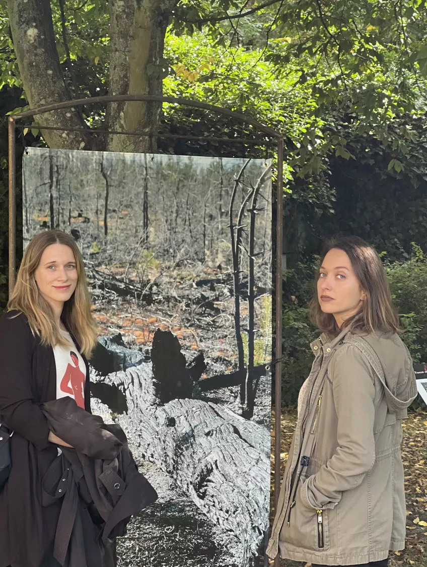 Lina and Pinar standing next to a huge photo of a charred log and burnt down forest in the aftermath of a fire. (Photo)